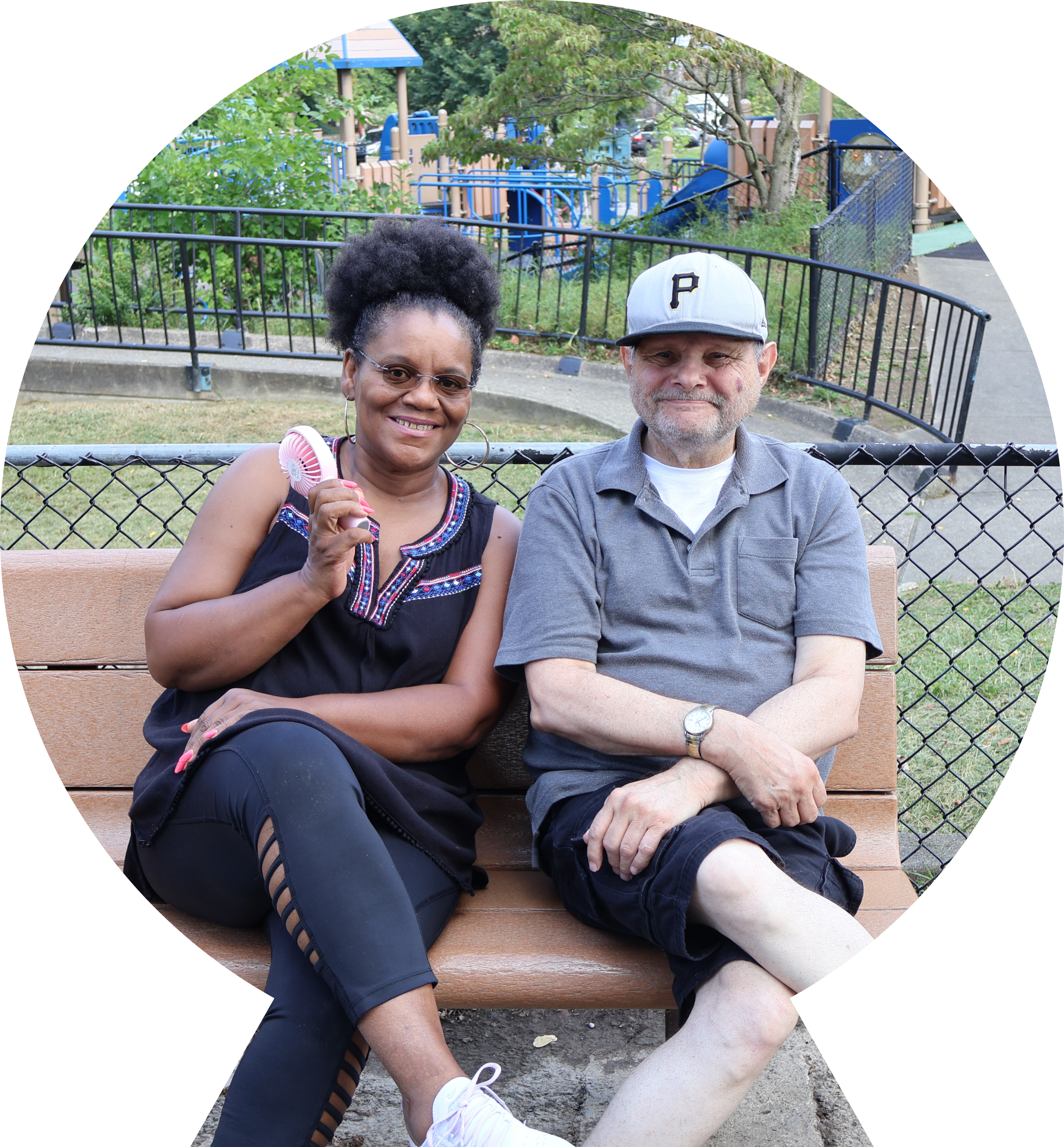 Woman with fan and man in a ball cap sit on a park bench smiling for the camera Woman with fan and man in a ball cap sit on a park bench smiling for the camera